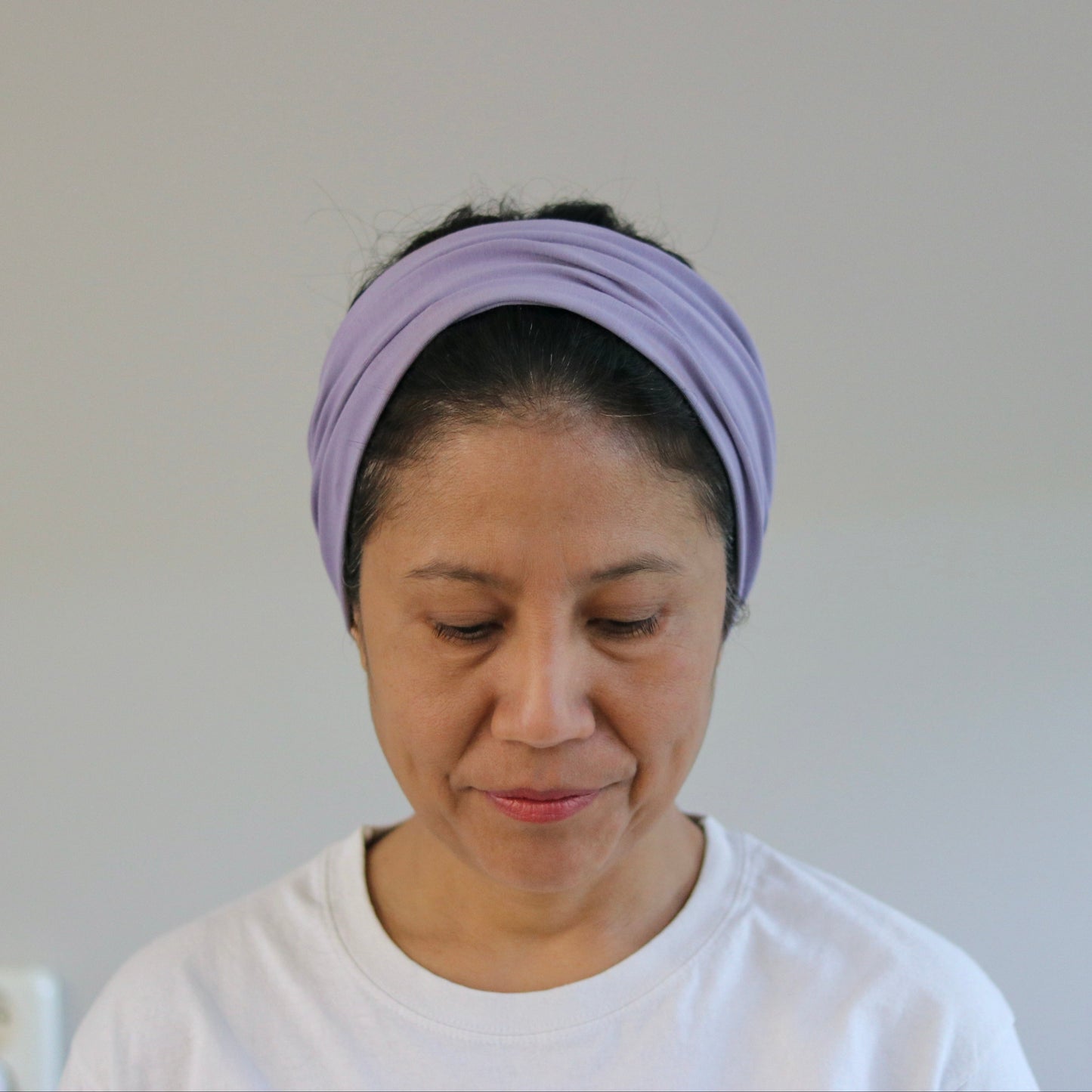 A woman wearing a light purple twisted headband and a white t-shirt, looking down against a plain light gray wall background.