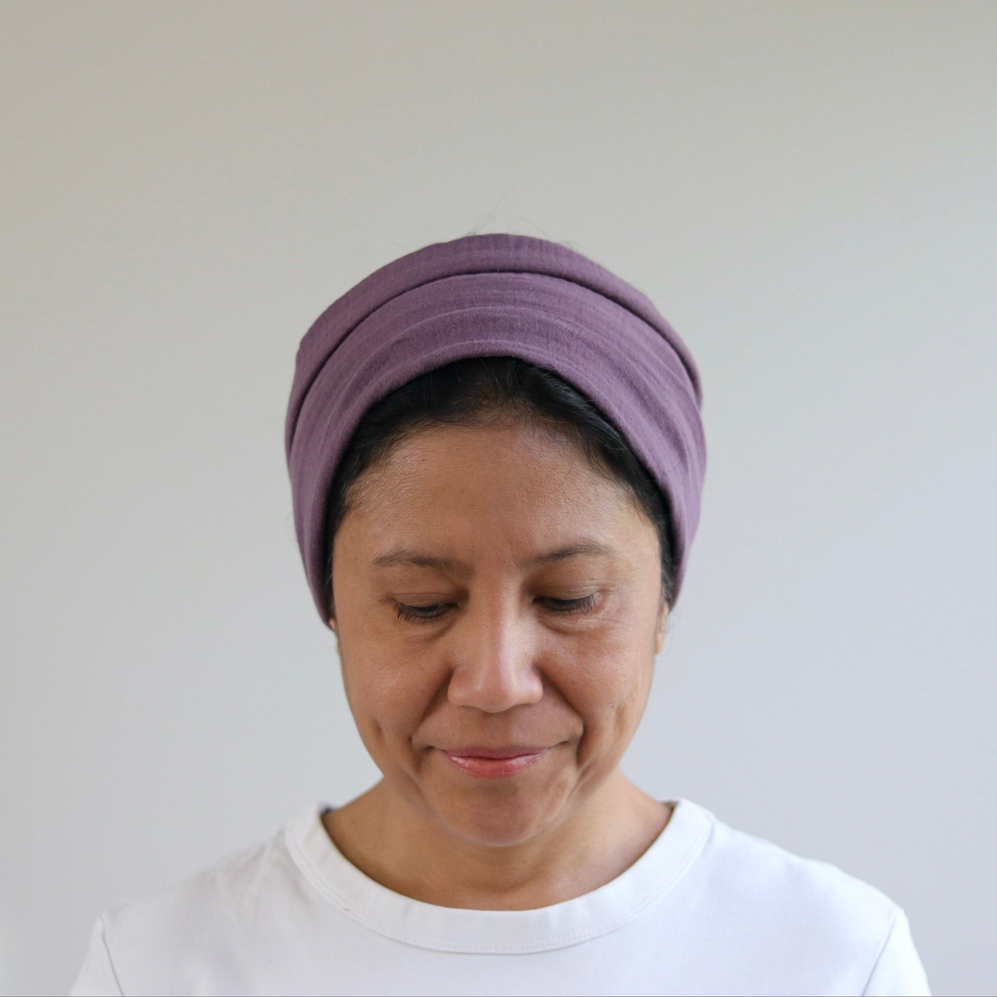 A woman wearing a mauve- purple fabric headwrap or turban-style hair accessory, paired with a white t-shirt, shown against a plain light gray wall.
