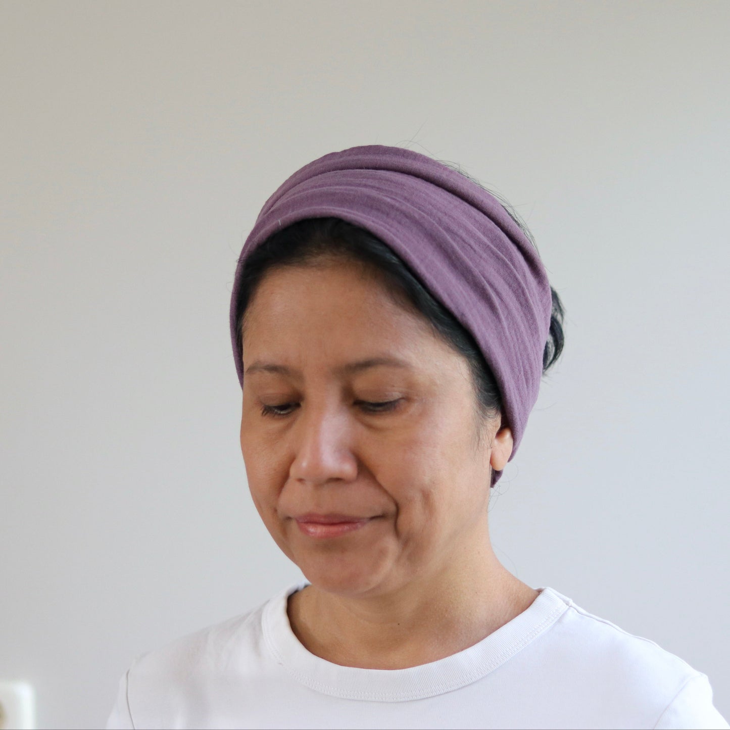 A woman wearing a mauve purple fabric headwrap or turban-style headband, paired with a white t-shirt, shown from the chest up against a plain light gray wall.
