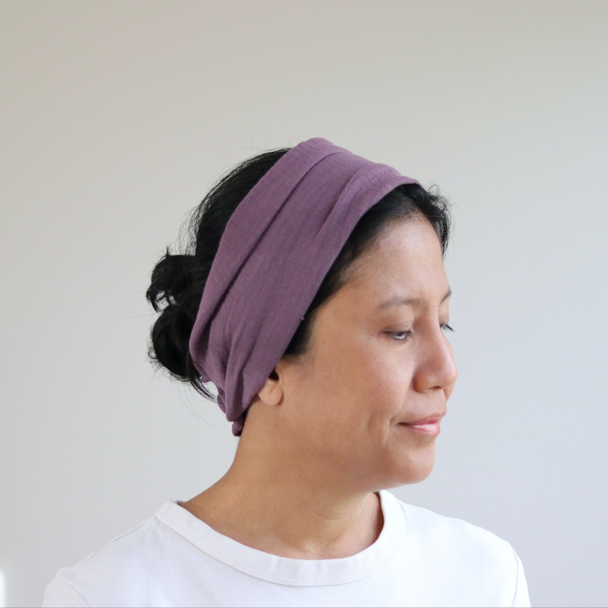 A woman wearing a wide, multi-layered mauve- purple fabric headband, shown in profile against a plain light gray wall.