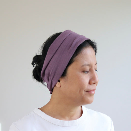 A woman wearing a wide, multi-layered mauve- purple fabric headband, shown in profile against a plain light gray wall.