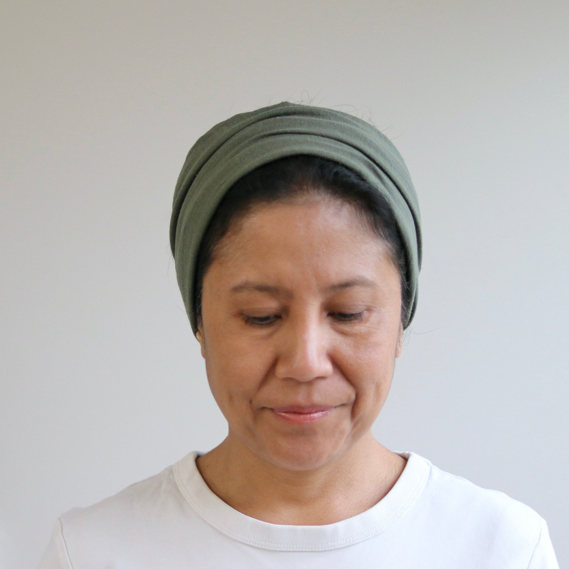 A woman wearing a dark olive green headwrap or turban-style cap, paired with a white crewneck top, standing against a plain light gray wall.