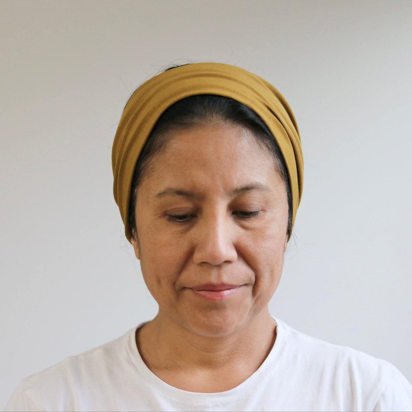 A woman wearing a mustard yellow headband and a white t-shirt, looking down with a neutral expression against a plain light gray wall.