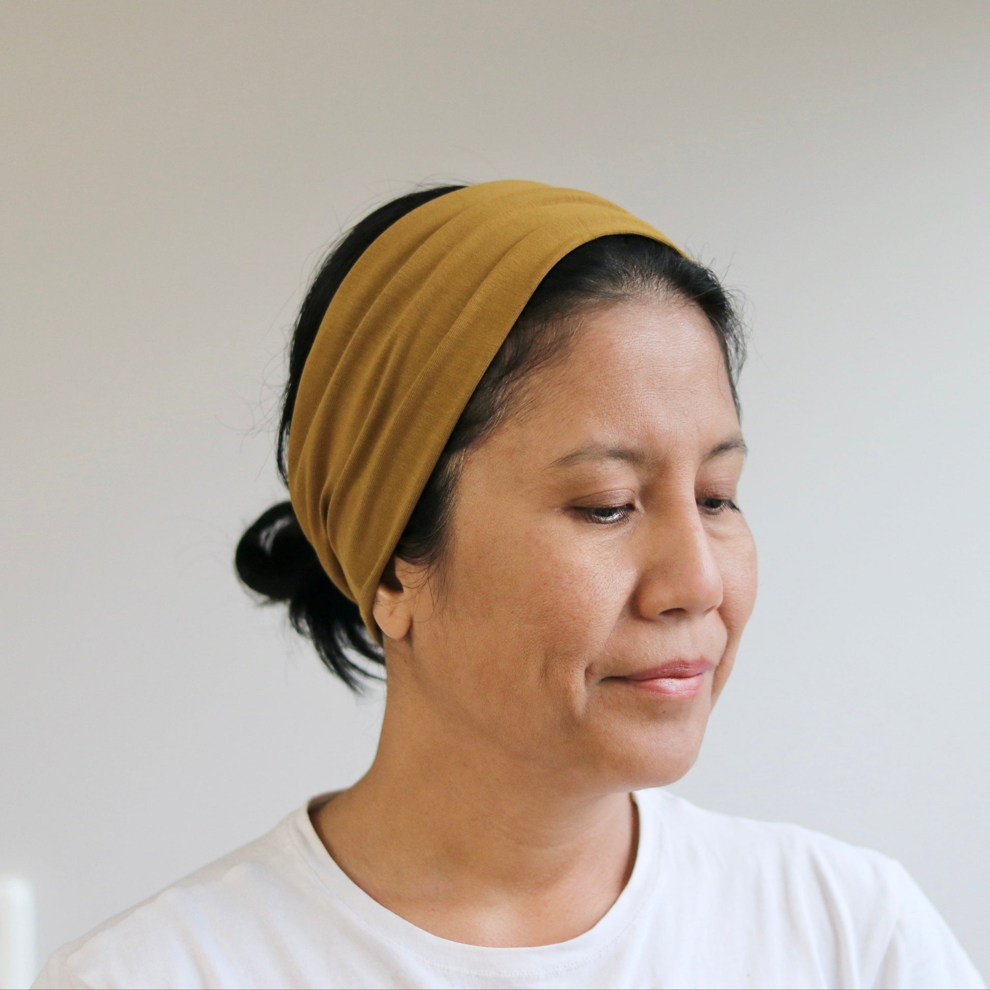 A woman wearing a mustard yellow fabric headband and a white t-shirt, shown in a close-up profile against a plain light gray wall.