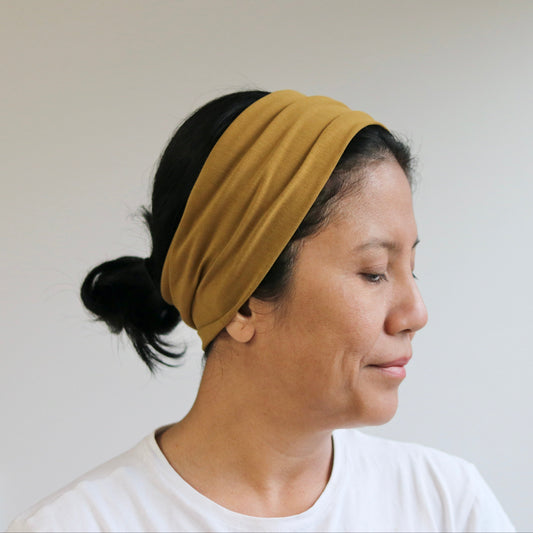 A woman wearing an ocher- brown fabric headband with her black hair tied back in a low bun, shown in profile against a plain light wall.