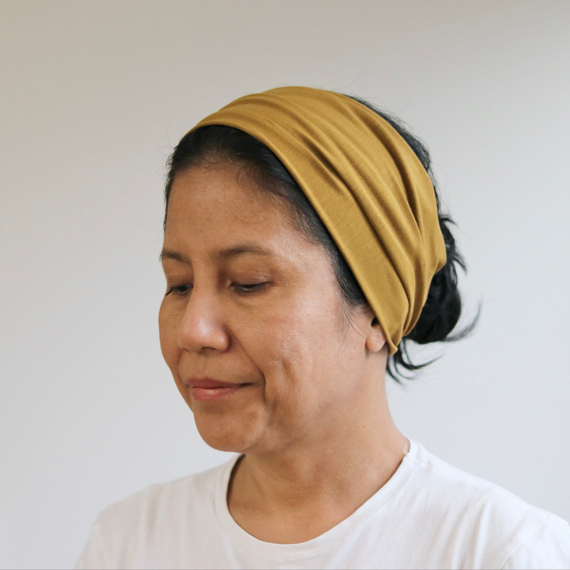A woman wearing a mustard yellow fabric headband and a white t-shirt, shown in a close-up profile view against a plain light gray wall.