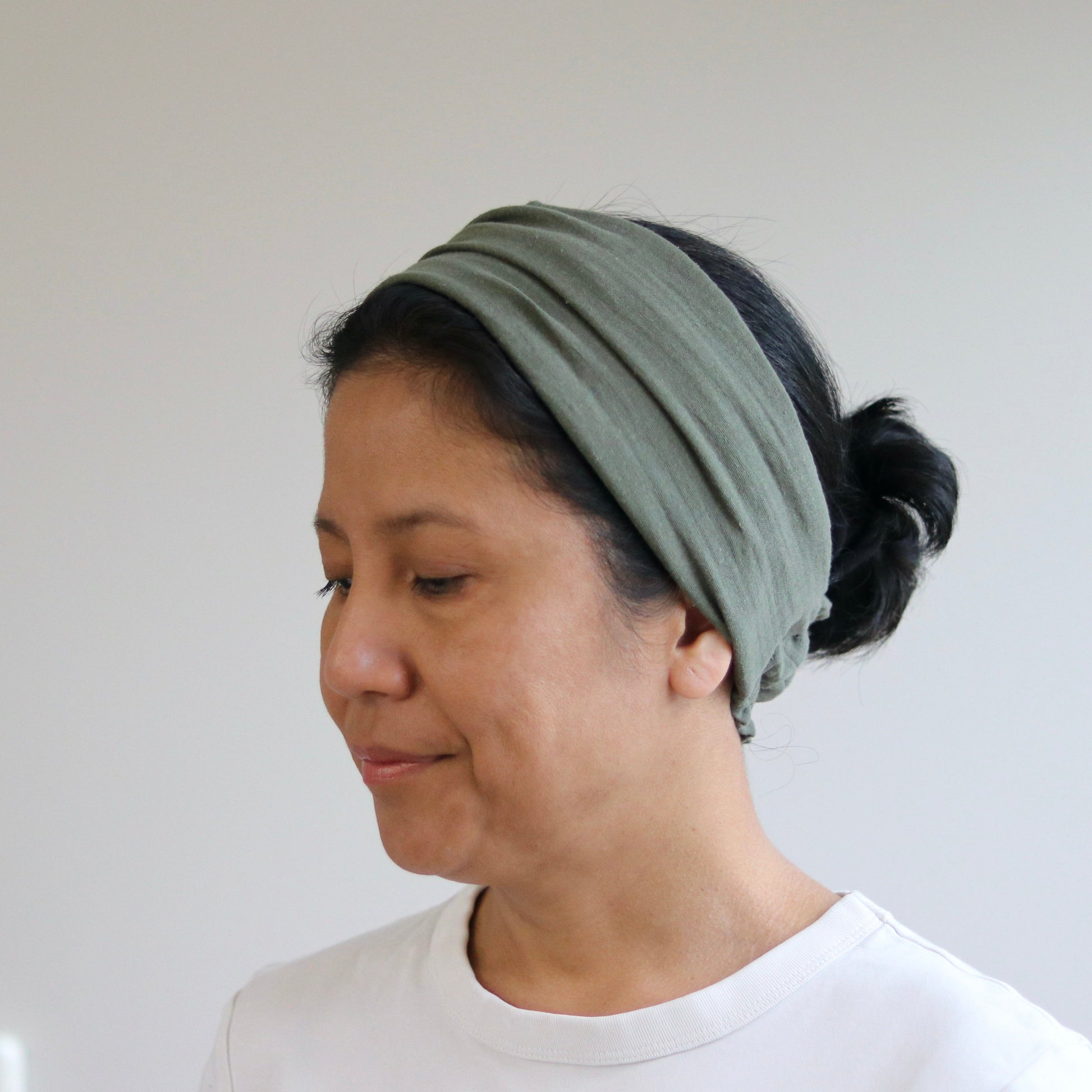 A woman wearing a wide, multi-layered sage green headband and a white t-shirt, shown in profile against a plain light gray wall.