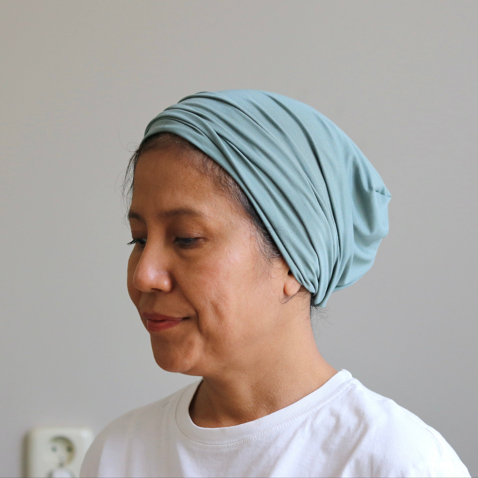 A woman wearing a light green pleated headscarf or hijab, paired with a white t-shirt, shown in profile against a plain light gray wall.
