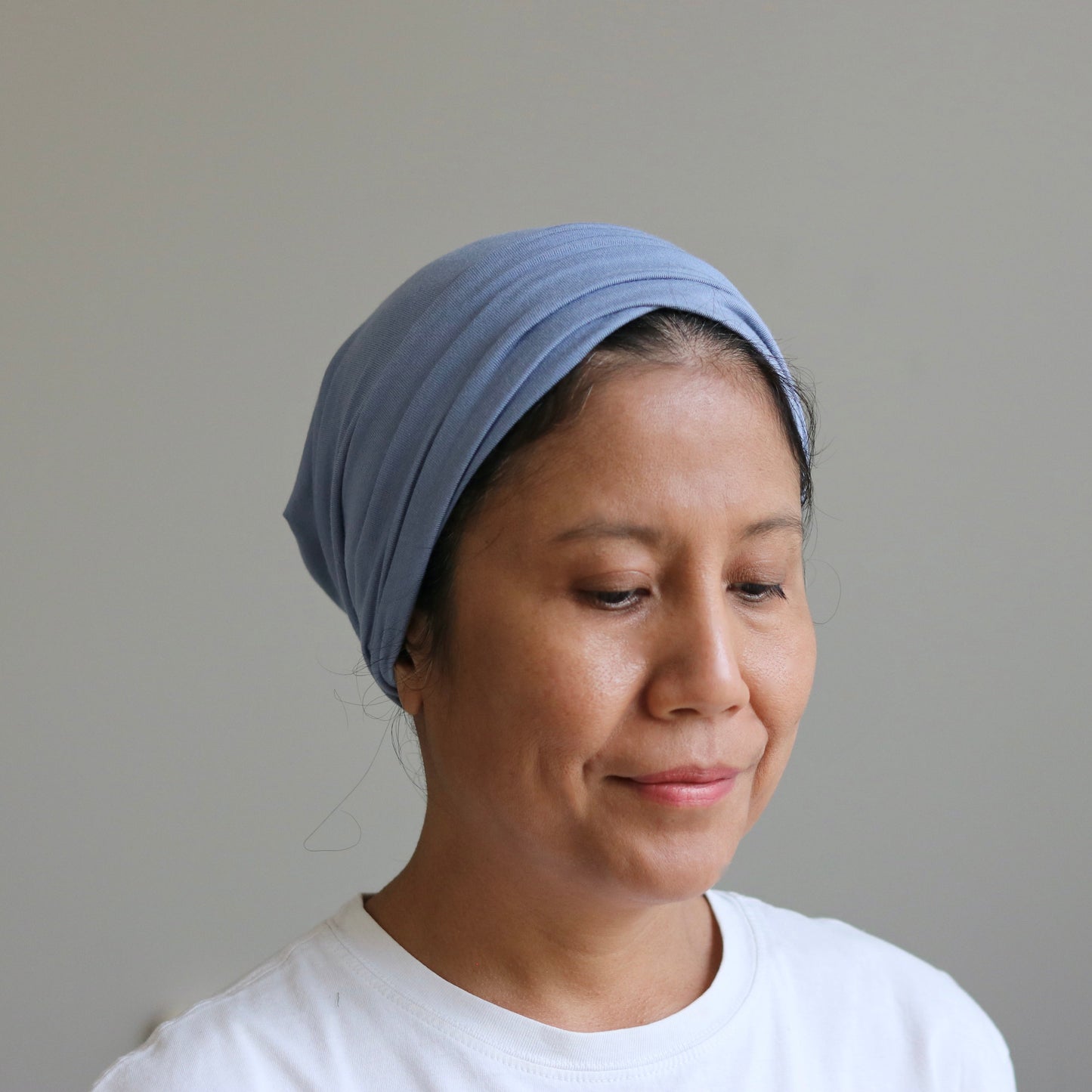 A woman wearing a light blue fabric headwrap or turban-style head covering, paired with a white t-shirt, shown against a plain light gray wall.