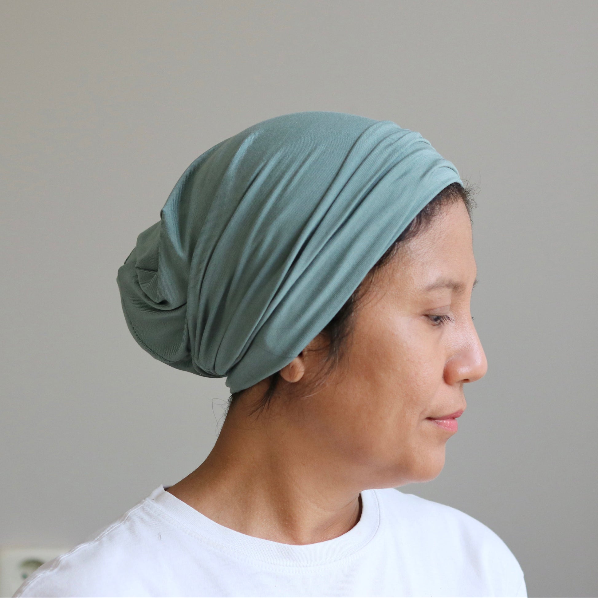 A woman in profile wearing a sage green knotted headscarf or hijab, paired with a white t-shirt, set against a plain light gray wall.