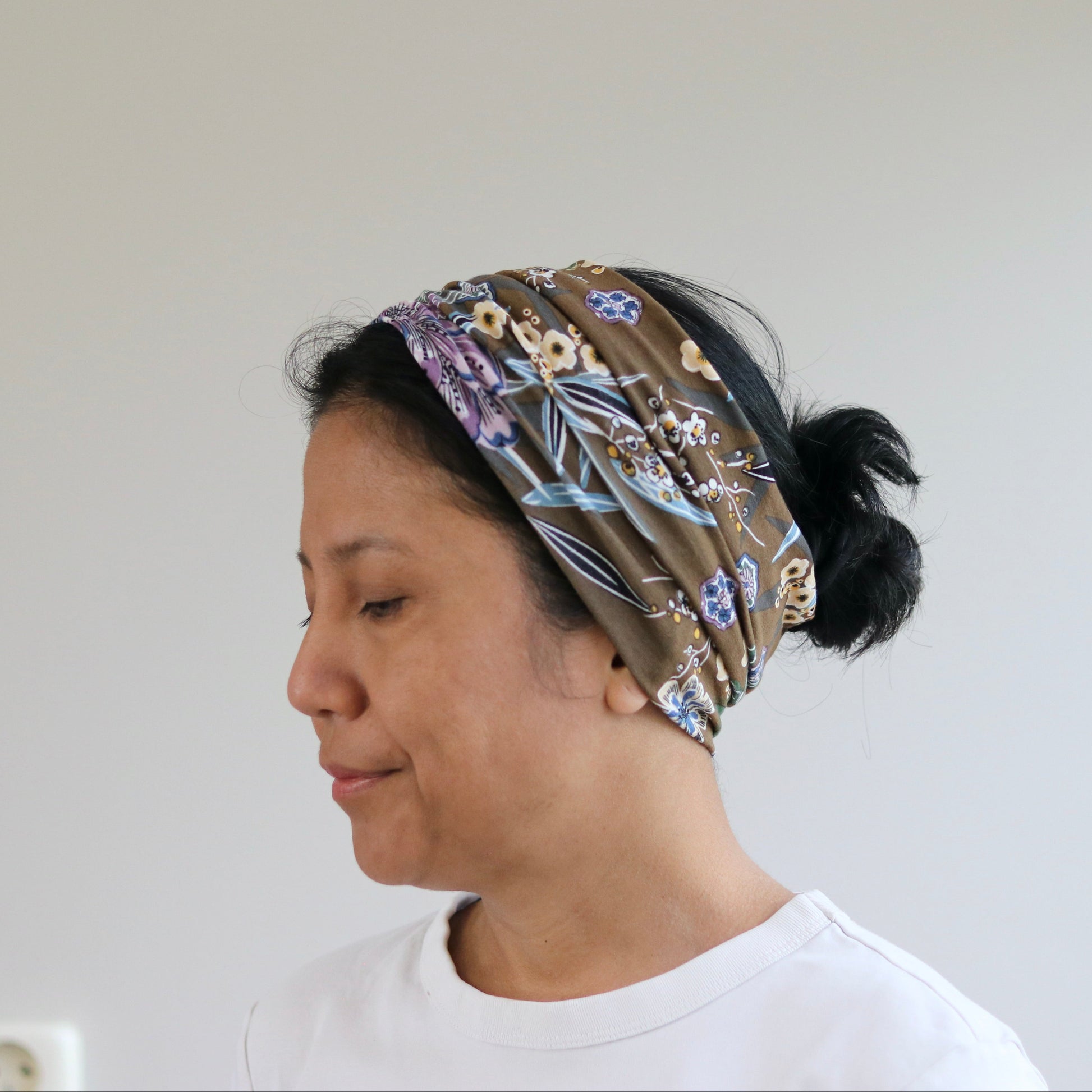 A woman in profile wearing a brown patterned headband with purple, blue, and beige floral details, paired with a white t-shirt against a plain light gray wall.