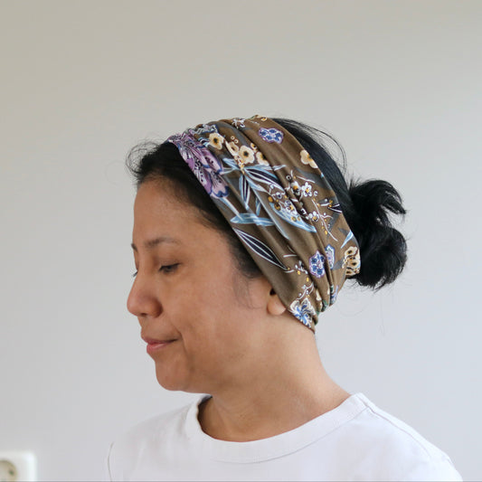 A woman in profile wearing a brown patterned headband with purple, blue, and beige floral details, paired with a white t-shirt against a plain light gray wall.