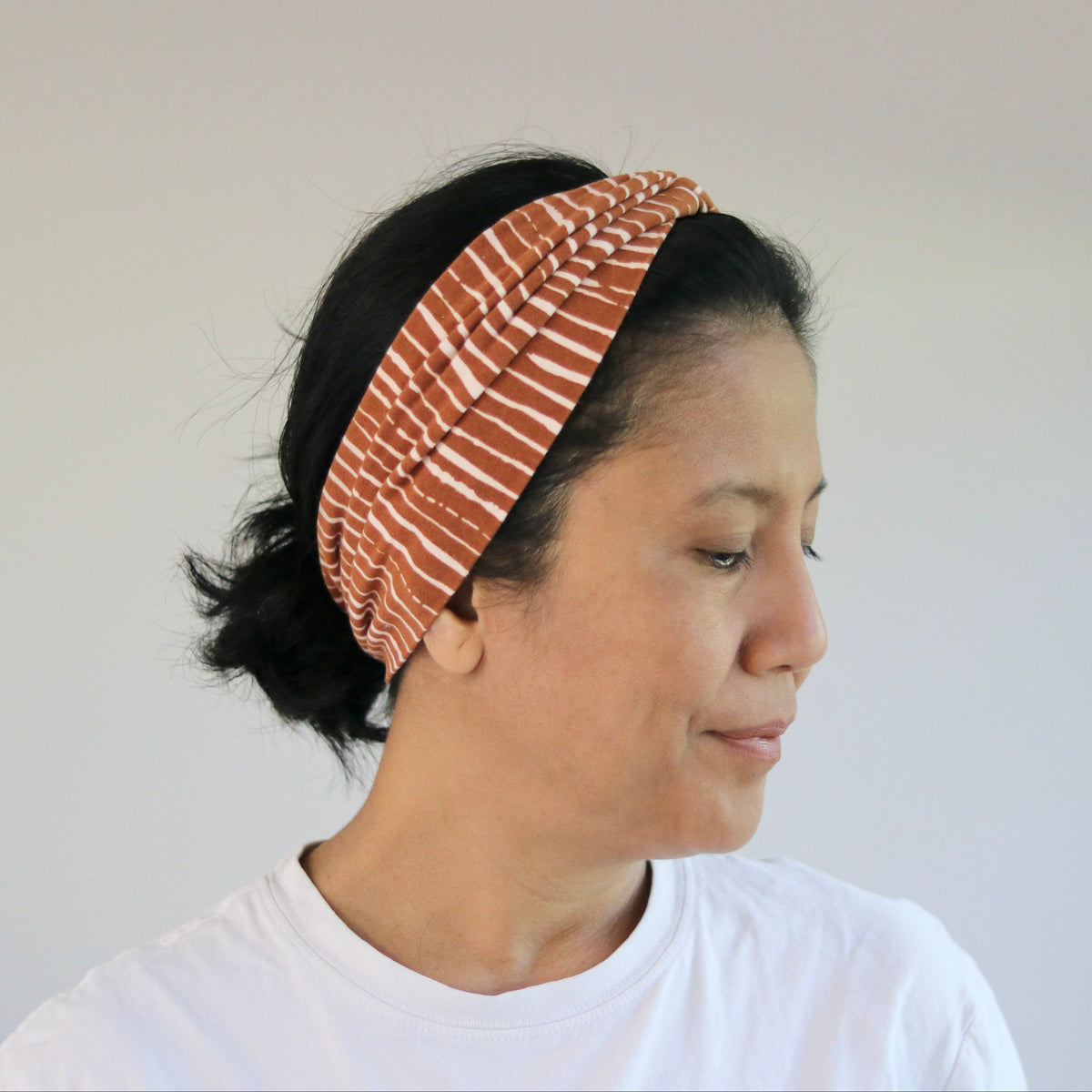 A woman wearing a wide, gathered headband with a rust-orange and white striped pattern, paired with a white t-shirt, shown in profile against a neutral indoor background.