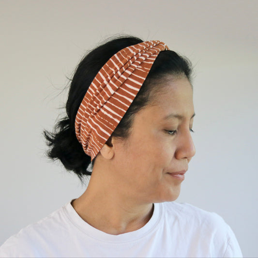 A woman wearing a wide, gathered headband with a rust-orange and white striped pattern, paired with a white t-shirt, shown in profile against a neutral indoor background.