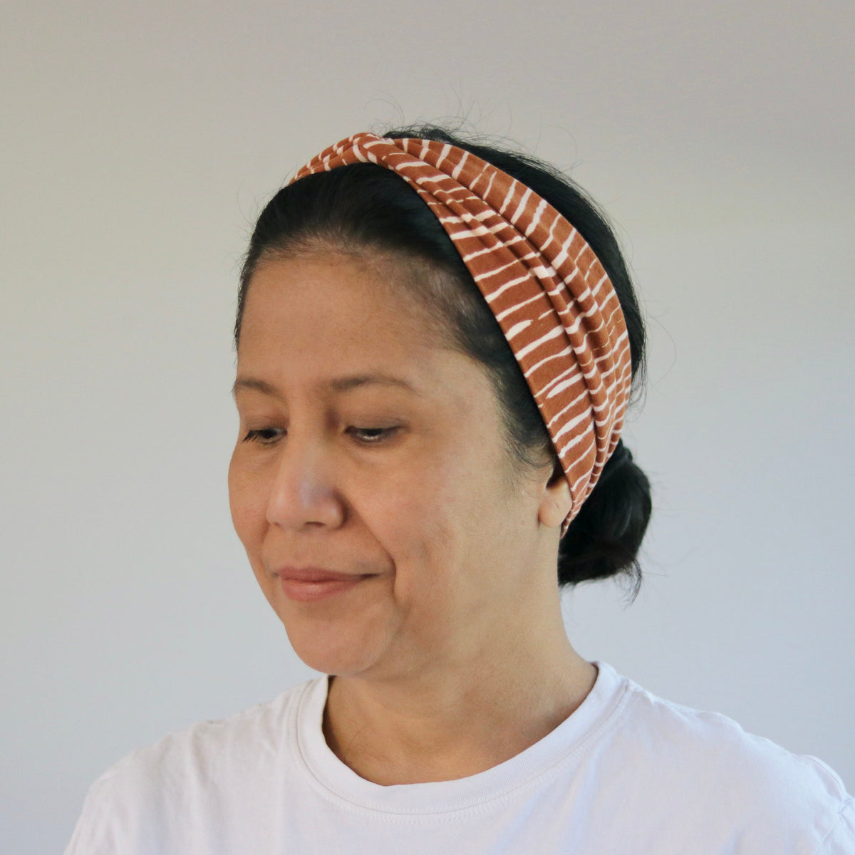 A woman wearing a rust-orange and white striped fabric headband, paired with a white t-shirt, shown in a brightly lit indoor setting.