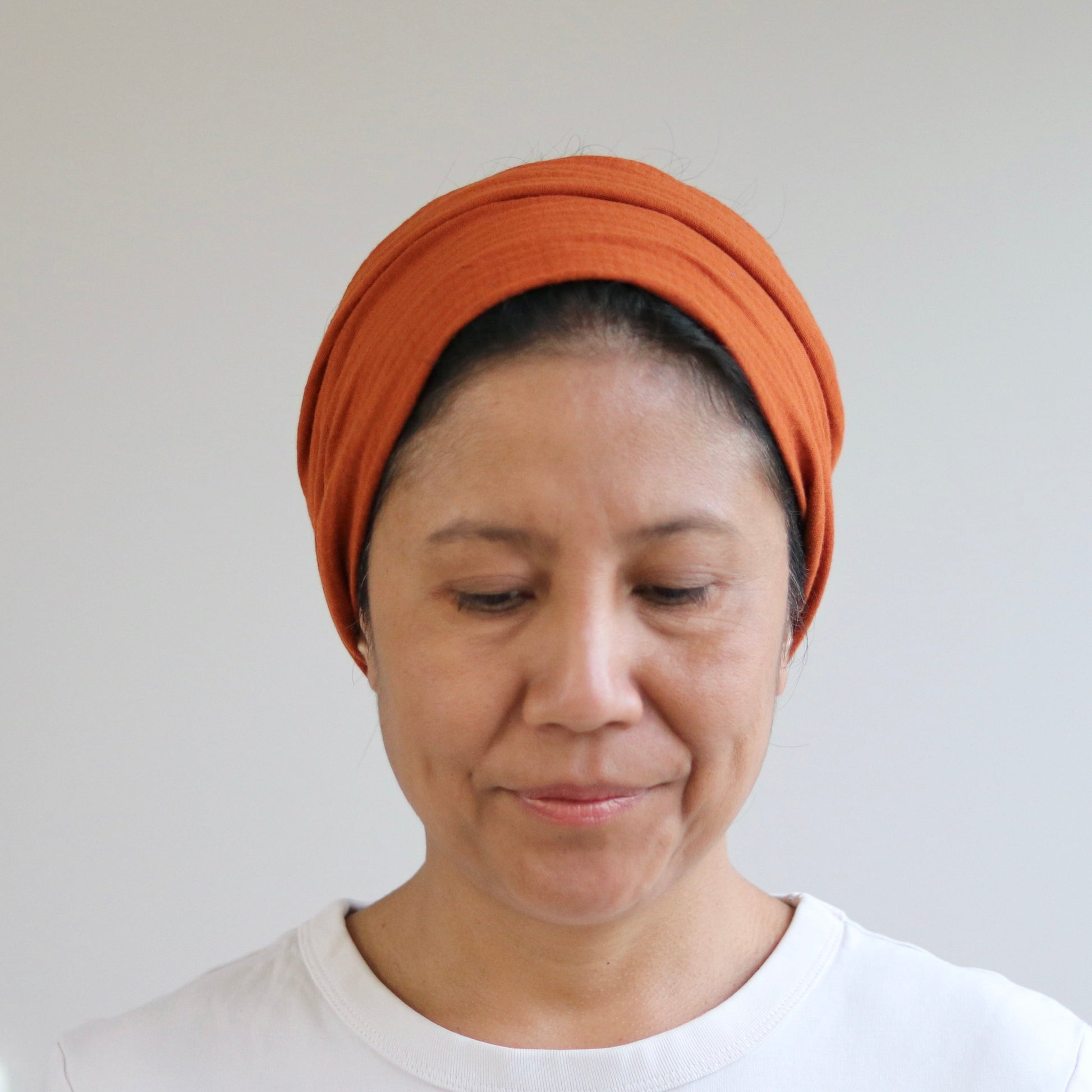 A woman wearing a terra muslin headwrap turban and a white t-shirt, looking down with a neutral expression against a plain light gray wall.