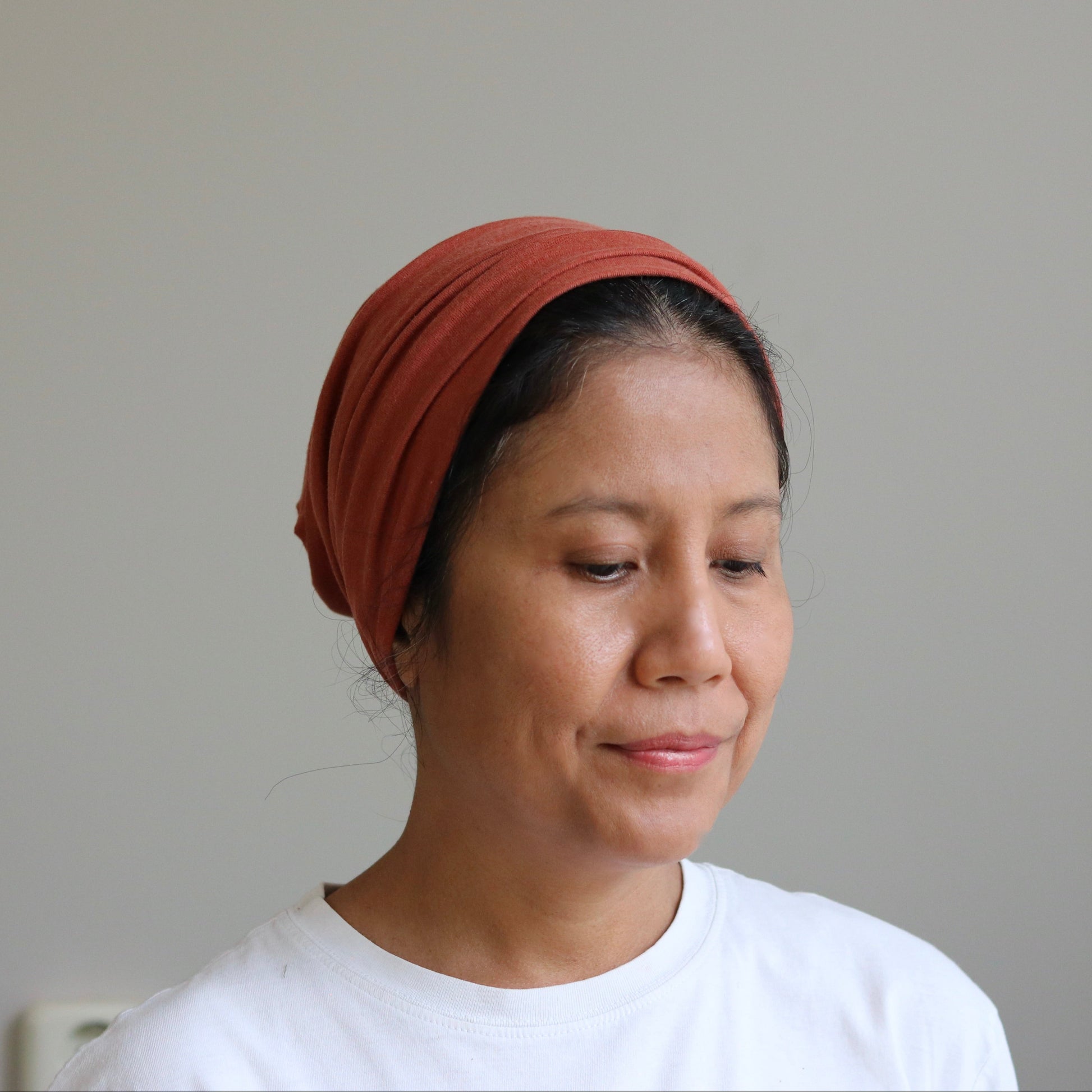 A woman wearing a rust-orange fabric headwrap or headband, paired with a white t-shirt, shown from the chest up against a plain light gray wall.