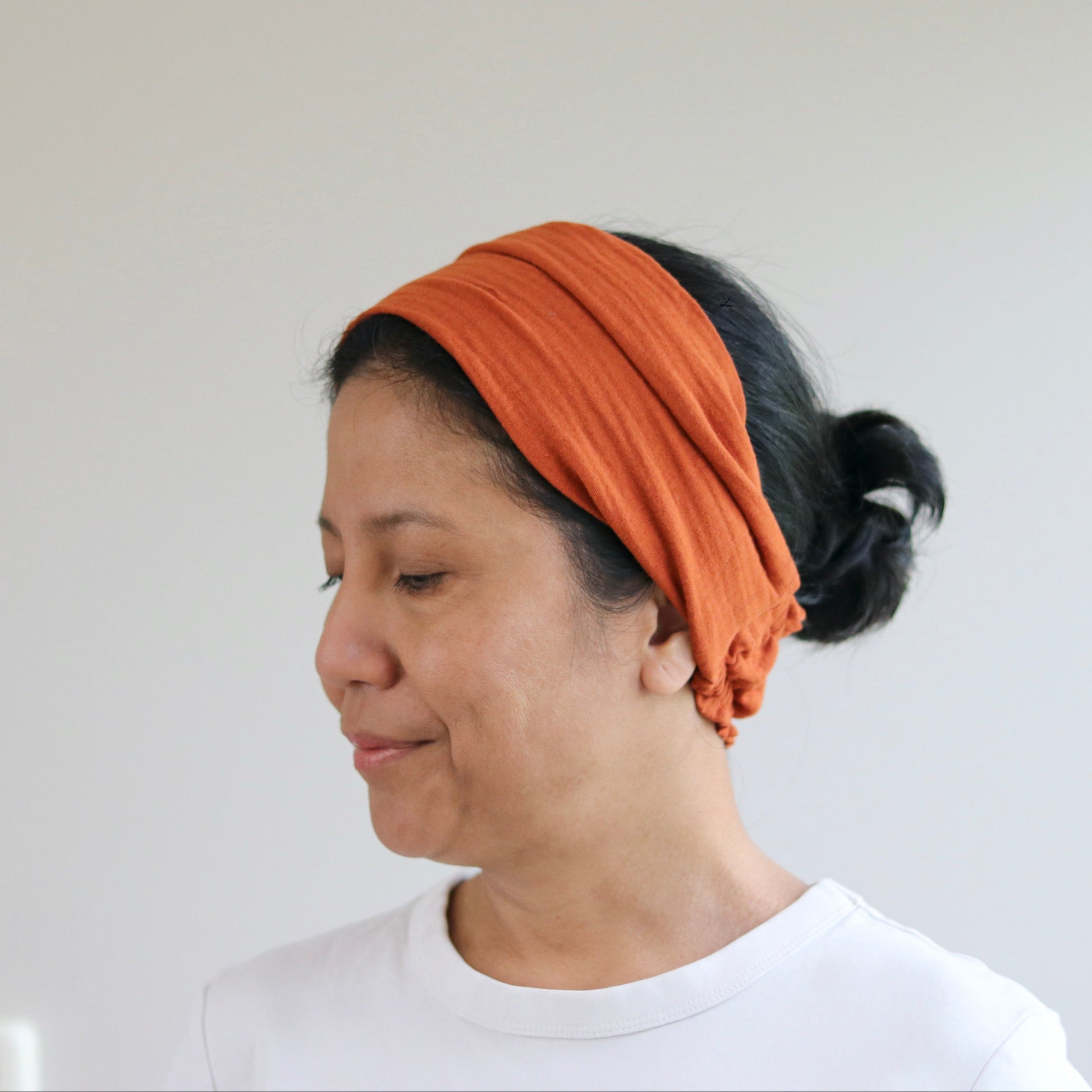 A woman wearing a soft, terracotta fabric headband and a white t-shirt, shown in profile against a plain light gray wall.