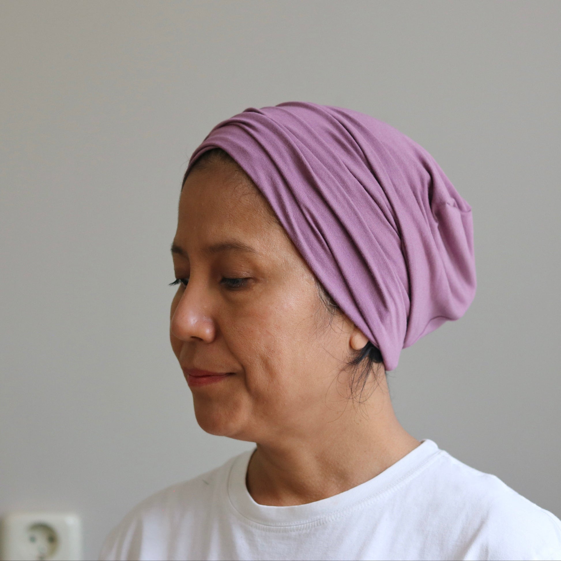 A woman wearing a soft lavender-purple knotted headscarf or turban-style cap, paired with a white t-shirt, shown in profile against a plain light gray wall.