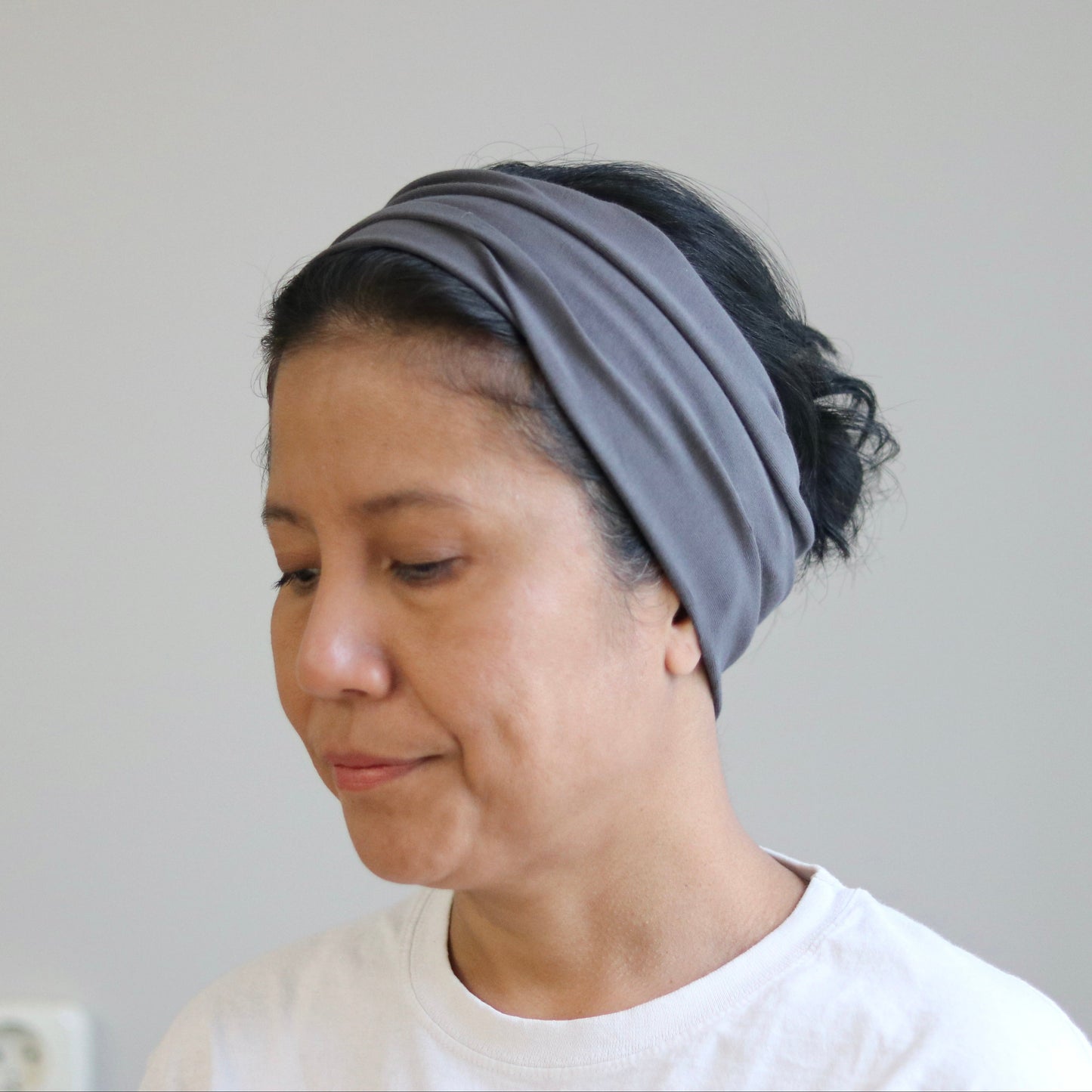 A woman wearing a gray fabric headband and a white t-shirt, looking down against a plain light gray wall.