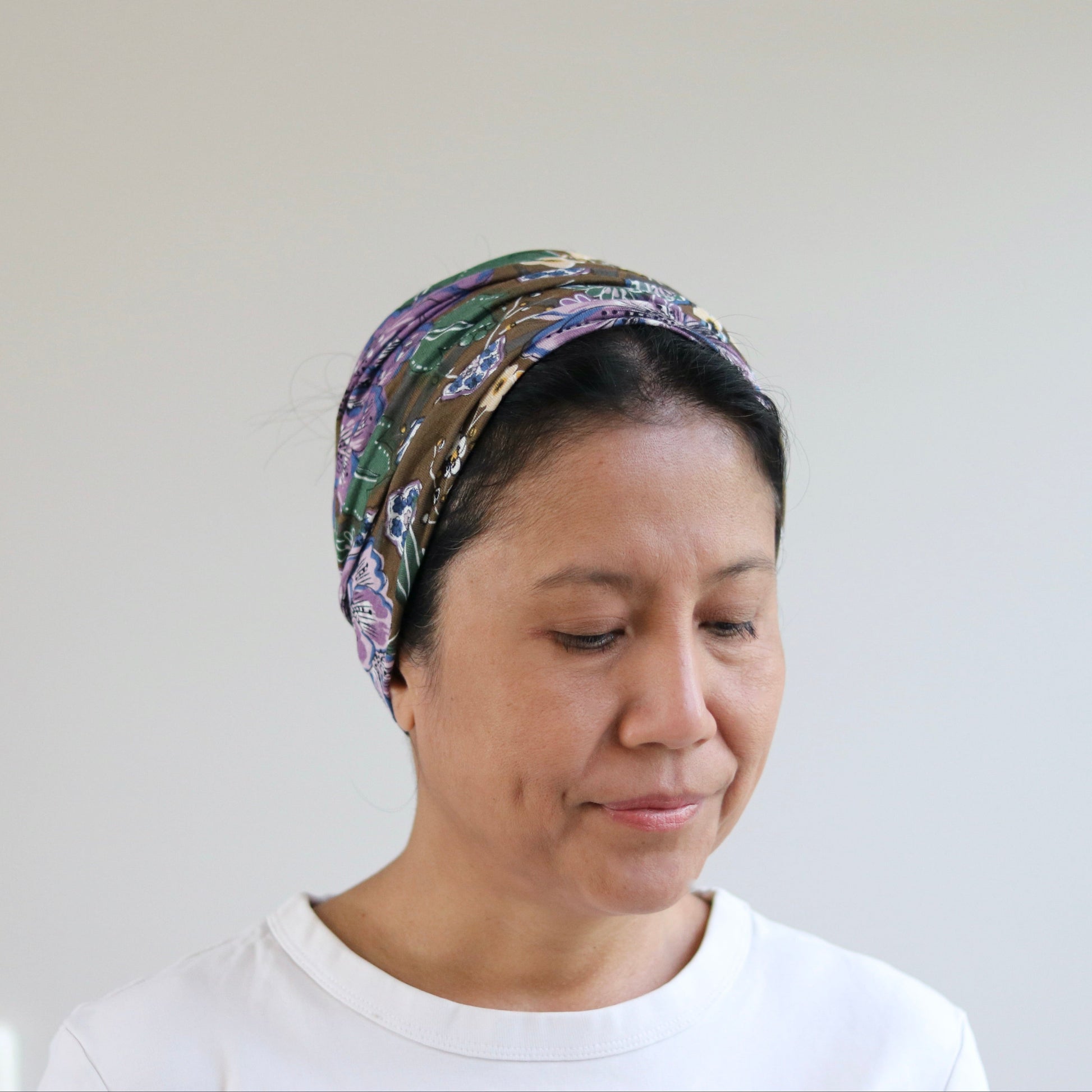 A woman wearing a patterned headwrap headband and a white t-shirt, looking down against a plain light gray wall background.