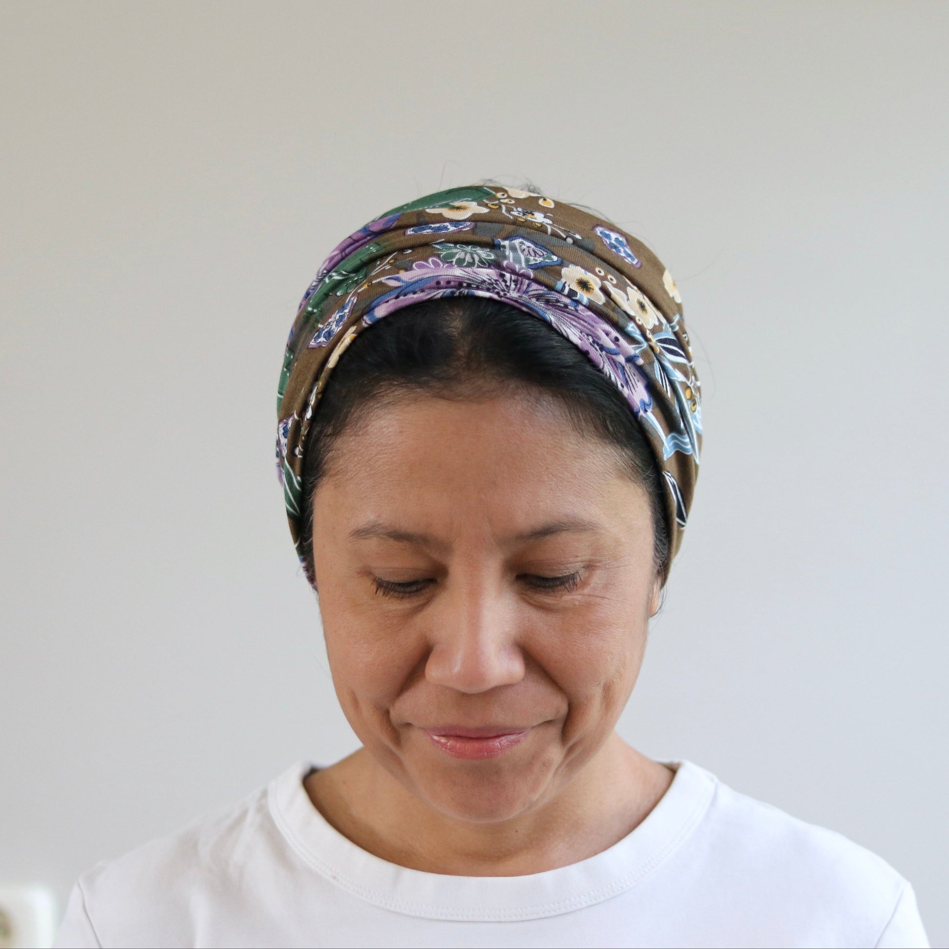 A woman wearing a patterned multicolor headscarf and a white t-shirt, looking down with a neutral expression against a plain light gray wall.